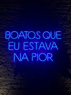 A neon sign displaying a phrase in Portuguese, illuminated in bright blue against a dark brick wall.