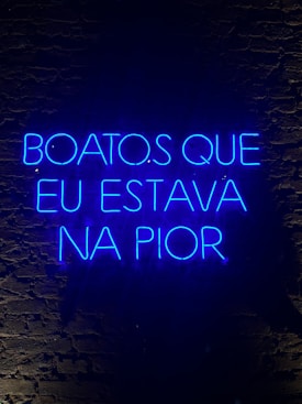 A neon sign displaying a phrase in Portuguese, illuminated in bright blue against a dark brick wall.