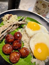 A colorful plate showcasing a traditional meal that includes two sunny-side-up eggs, grilled dried fish, savory meatballs, and a portion of shredded vegetable garnish. The foods are presented on a bed of white rice, served over a banana leaf, accompanied by a mug with the word 'Hola!' printed on it, and a beverage in the background.