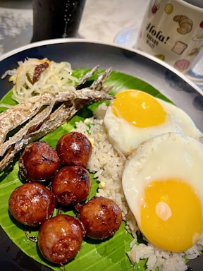 A colorful plate showcasing a traditional meal that includes two sunny-side-up eggs, grilled dried fish, savory meatballs, and a portion of shredded vegetable garnish. The foods are presented on a bed of white rice, served over a banana leaf, accompanied by a mug with the word 'Hola!' printed on it, and a beverage in the background.