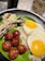 A colorful plate showcasing a traditional meal that includes two sunny-side-up eggs, grilled dried fish, savory meatballs, and a portion of shredded vegetable garnish. The foods are presented on a bed of white rice, served over a banana leaf, accompanied by a mug with the word 'Hola!' printed on it, and a beverage in the background.