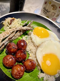 A colorful plate showcasing a traditional meal that includes two sunny-side-up eggs, grilled dried fish, savory meatballs, and a portion of shredded vegetable garnish. The foods are presented on a bed of white rice, served over a banana leaf, accompanied by a mug with the word 'Hola!' printed on it, and a beverage in the background.
