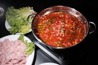 A hot pot filled with a red-colored broth containing various vegetables such as peppers and garnished with sesame seeds is placed on a black stovetop. Surrounding the pot are plates with fresh lettuce leaves and raw sliced meat, ready to be cooked.