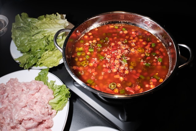 A hot pot filled with a red-colored broth containing various vegetables such as peppers and garnished with sesame seeds is placed on a black stovetop. Surrounding the pot are plates with fresh lettuce leaves and raw sliced meat, ready to be cooked.