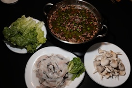 The image displays a table with a hot pot containing broth filled with sliced green peppers and seeds. Surrounding the hot pot are three plates, one with fresh green lettuce, another with sliced raw fish, and the third with a pile of uncooked mushrooms.