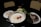 A hot pot with a creamy white broth contains red dates and ginseng. Surrounding it are plates of mushrooms, leafy greens, and sliced meat on a dark table with woven back chairs in the background.