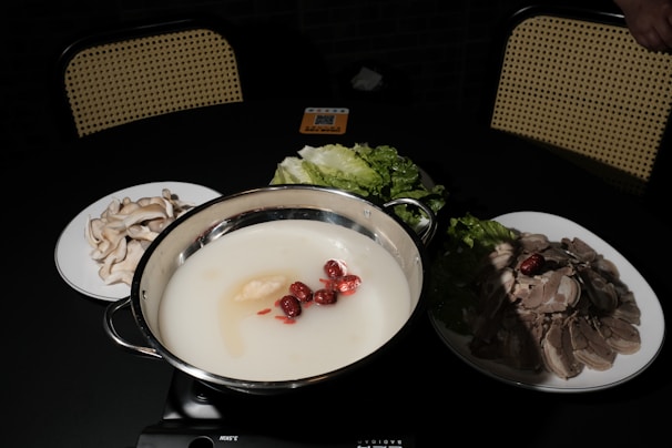 A hot pot with a creamy white broth contains red dates and ginseng. Surrounding it are plates of mushrooms, leafy greens, and sliced meat on a dark table with woven back chairs in the background.