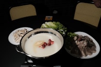 A hot pot with a creamy white broth contains red dates and ginseng. Surrounding it are plates of mushrooms, leafy greens, and sliced meat on a dark table with woven back chairs in the background.