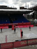 A beach volleyball court is set up in an outdoor arena, with four individuals engaging in a casual play or practice session. The court is surrounded by red banners and a seating area with rows of blue chairs. A large industrial structure can be seen in the background, contrasting with the sporting event. An umbrella bearing a logo provides shade at one end of the court, and volleyball equipment is visible.