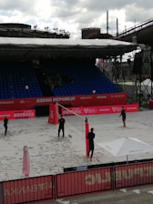 A beach volleyball court is set up in an outdoor arena, with four individuals engaging in a casual play or practice session. The court is surrounded by red banners and a seating area with rows of blue chairs. A large industrial structure can be seen in the background, contrasting with the sporting event. An umbrella bearing a logo provides shade at one end of the court, and volleyball equipment is visible.