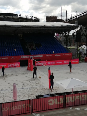 A beach volleyball court is set up in an outdoor arena, with four individuals engaging in a casual play or practice session. The court is surrounded by red banners and a seating area with rows of blue chairs. A large industrial structure can be seen in the background, contrasting with the sporting event. An umbrella bearing a logo provides shade at one end of the court, and volleyball equipment is visible.