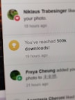 A digital notification on a screen announces the achievement of reaching 500,000 downloads. The message is accompanied by a gold trophy icon symbolizing success. The background shows other notifications with interactions from a social media platform.