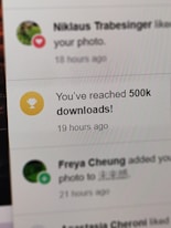 A digital notification on a screen announces the achievement of reaching 500,000 downloads. The message is accompanied by a gold trophy icon symbolizing success. The background shows other notifications with interactions from a social media platform.