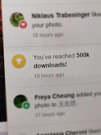 A digital notification on a screen announces the achievement of reaching 500,000 downloads. The message is accompanied by a gold trophy icon symbolizing success. The background shows other notifications with interactions from a social media platform.
