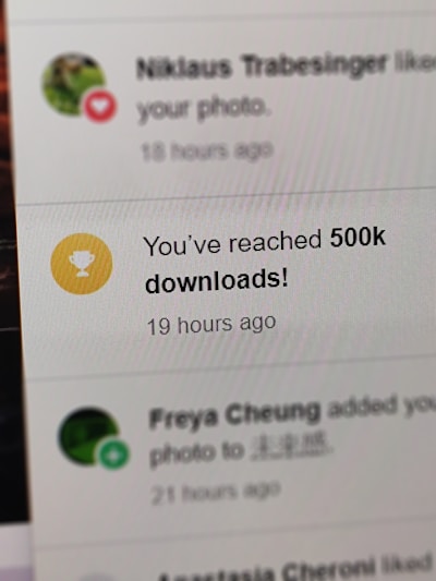 A digital notification on a screen announces the achievement of reaching 500,000 downloads. The message is accompanied by a gold trophy icon symbolizing success. The background shows other notifications with interactions from a social media platform.