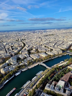 Aerial view of a city with a dense layout of traditional European architecture, with the Seine River curving through the cityscape. Bridges connect the two riverbanks, with several boats visible on the water. Patches of green spaces and parks are interspersed among the buildings. The sky is clear with a few scattered clouds.