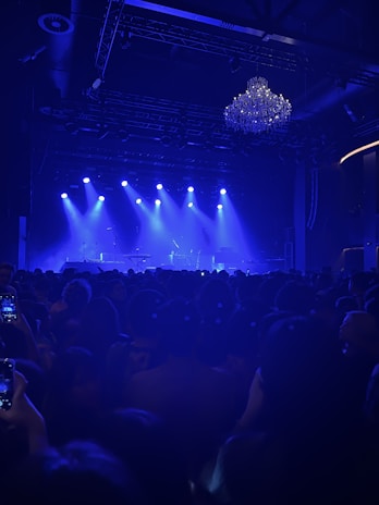 A concert venue filled with people, all facing a stage that is illuminated by blue lights. On the stage, microphones and musical equipment can be seen, set up for a performance. A large, ornate chandelier hangs from the ceiling, adding to the atmosphere.