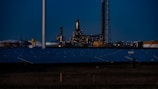 A nighttime industrial scene featuring a large factory with numerous lights illuminating its structure. Tall chimneys and poles are prominent against a dark sky, while rows of solar panels spread across the foreground. The overall setting appears to be a mix of industrial and renewable energy components.