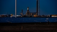A nighttime industrial scene featuring a large factory with numerous lights illuminating its structure. Tall chimneys and poles are prominent against a dark sky, while rows of solar panels spread across the foreground. The overall setting appears to be a mix of industrial and renewable energy components.