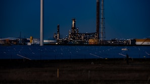 A nighttime industrial scene featuring a large factory with numerous lights illuminating its structure. Tall chimneys and poles are prominent against a dark sky, while rows of solar panels spread across the foreground. The overall setting appears to be a mix of industrial and renewable energy components.