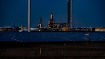 A nighttime industrial scene featuring a large factory with numerous lights illuminating its structure. Tall chimneys and poles are prominent against a dark sky, while rows of solar panels spread across the foreground. The overall setting appears to be a mix of industrial and renewable energy components.