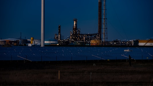 A nighttime industrial scene featuring a large factory with numerous lights illuminating its structure. Tall chimneys and poles are prominent against a dark sky, while rows of solar panels spread across the foreground. The overall setting appears to be a mix of industrial and renewable energy components.