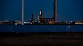 A nighttime industrial scene featuring a large factory with numerous lights illuminating its structure. Tall chimneys and poles are prominent against a dark sky, while rows of solar panels spread across the foreground. The overall setting appears to be a mix of industrial and renewable energy components.