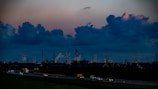 The image depicts a sprawling industrial landscape with several smokestacks and wind turbines visible against a dramatic evening sky filled with large, dark clouds. The foreground features a highway with vehicles moving in both directions, casting blurred lights, while the background is dominated by the industrial structures.