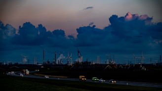 The image depicts a sprawling industrial landscape with several smokestacks and wind turbines visible against a dramatic evening sky filled with large, dark clouds. The foreground features a highway with vehicles moving in both directions, casting blurred lights, while the background is dominated by the industrial structures.