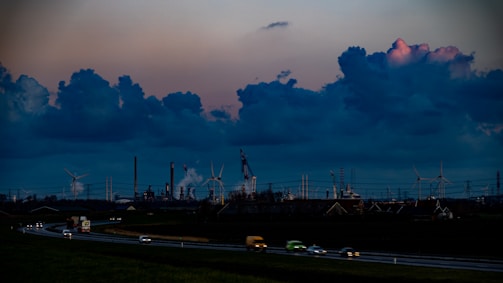 The image depicts a sprawling industrial landscape with several smokestacks and wind turbines visible against a dramatic evening sky filled with large, dark clouds. The foreground features a highway with vehicles moving in both directions, casting blurred lights, while the background is dominated by the industrial structures.