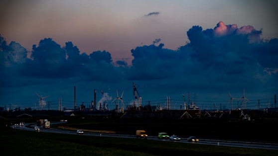 The image depicts a sprawling industrial landscape with several smokestacks and wind turbines visible against a dramatic evening sky filled with large, dark clouds. The foreground features a highway with vehicles moving in both directions, casting blurred lights, while the background is dominated by the industrial structures.