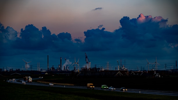 The image depicts a sprawling industrial landscape with several smokestacks and wind turbines visible against a dramatic evening sky filled with large, dark clouds. The foreground features a highway with vehicles moving in both directions, casting blurred lights, while the background is dominated by the industrial structures.