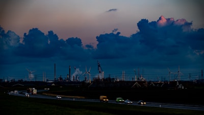 The image depicts a sprawling industrial landscape with several smokestacks and wind turbines visible against a dramatic evening sky filled with large, dark clouds. The foreground features a highway with vehicles moving in both directions, casting blurred lights, while the background is dominated by the industrial structures.