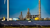 A landscape featuring a large solar panel array in the foreground, with a modern wind turbine to the left. Behind the renewable energy installations, an industrial facility with multiple chimneys and storage tanks is visible under a clear blue sky.