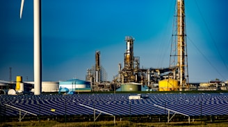 A landscape featuring a large solar panel array in the foreground, with a modern wind turbine to the left. Behind the renewable energy installations, an industrial facility with multiple chimneys and storage tanks is visible under a clear blue sky.