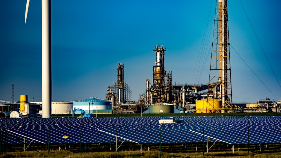 A landscape featuring a large solar panel array in the foreground, with a modern wind turbine to the left. Behind the renewable energy installations, an industrial facility with multiple chimneys and storage tanks is visible under a clear blue sky.