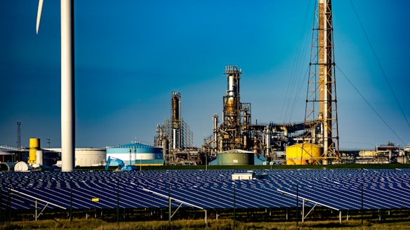 A landscape featuring a large solar panel array in the foreground, with a modern wind turbine to the left. Behind the renewable energy installations, an industrial facility with multiple chimneys and storage tanks is visible under a clear blue sky.