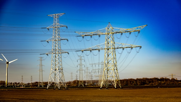A landscape with multiple electricity transmission towers and power lines stretching across a flat expanse. A wind turbine is visible to the left side of the image. The sky is clear and bright blue, suggesting a sunny day. In the distance, a line of trees marks the boundary of the open field.