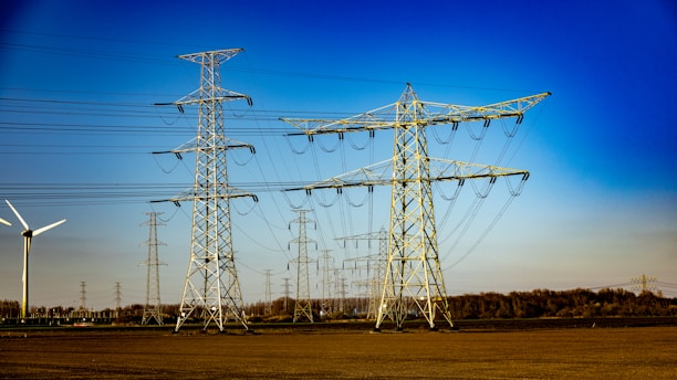 A landscape with multiple electricity transmission towers and power lines stretching across a flat expanse. A wind turbine is visible to the left side of the image. The sky is clear and bright blue, suggesting a sunny day. In the distance, a line of trees marks the boundary of the open field.