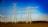 A landscape with multiple electricity transmission towers and power lines stretching across a flat expanse. A wind turbine is visible to the left side of the image. The sky is clear and bright blue, suggesting a sunny day. In the distance, a line of trees marks the boundary of the open field.