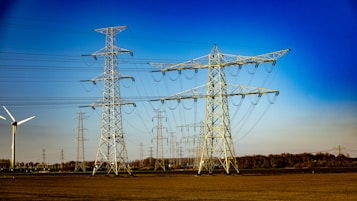 A landscape with multiple electricity transmission towers and power lines stretching across a flat expanse. A wind turbine is visible to the left side of the image. The sky is clear and bright blue, suggesting a sunny day. In the distance, a line of trees marks the boundary of the open field.