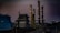 A complex industrial facility featuring tall, illuminated structures resembling chimneys or distillation columns. The scene is set during twilight, accentuated by gentle artificial lighting against a dim sky. Surrounding elements include large tanks and a perimeter fence, with piles of construction material visible.