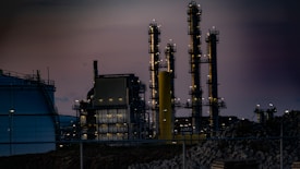 A complex industrial facility featuring tall, illuminated structures resembling chimneys or distillation columns. The scene is set during twilight, accentuated by gentle artificial lighting against a dim sky. Surrounding elements include large tanks and a perimeter fence, with piles of construction material visible.