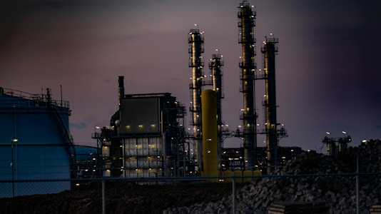 A complex industrial facility featuring tall, illuminated structures resembling chimneys or distillation columns. The scene is set during twilight, accentuated by gentle artificial lighting against a dim sky. Surrounding elements include large tanks and a perimeter fence, with piles of construction material visible.