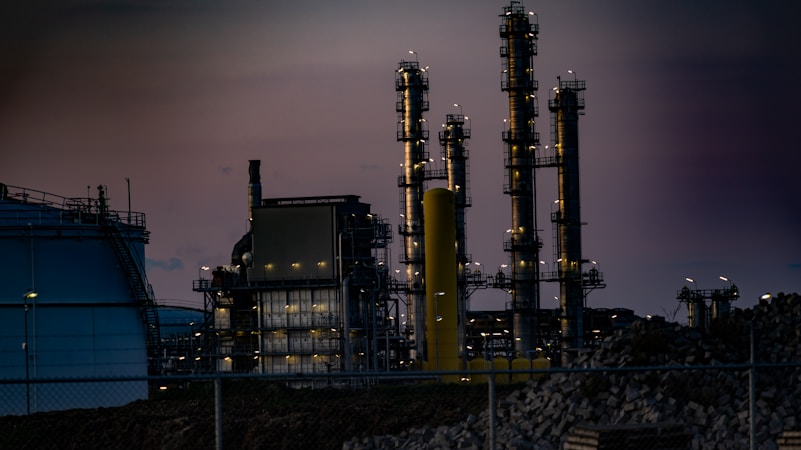 A complex industrial facility featuring tall, illuminated structures resembling chimneys or distillation columns. The scene is set during twilight, accentuated by gentle artificial lighting against a dim sky. Surrounding elements include large tanks and a perimeter fence, with piles of construction material visible.