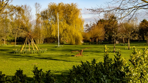 A sprawling garden landscape features lush green grass, bare trees, and a weeping willow. Wooden structures and an antique plow are situated on the lawn, surrounded by flower beds of budding plants ready for spring.