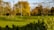 A sprawling garden landscape features lush green grass, bare trees, and a weeping willow. Wooden structures and an antique plow are situated on the lawn, surrounded by flower beds of budding plants ready for spring.
