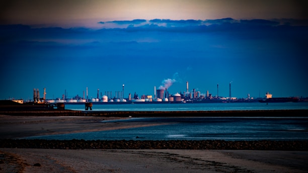 An industrial landscape featuring numerous large cylindrical tanks and distillation columns in the background, likely part of a refinery or chemical plant. Smoke is visible rising from some structures, indicating active industrial processes. The foreground includes a sandy shoreline with calm waters reflecting the blue and gray tones of the sky.