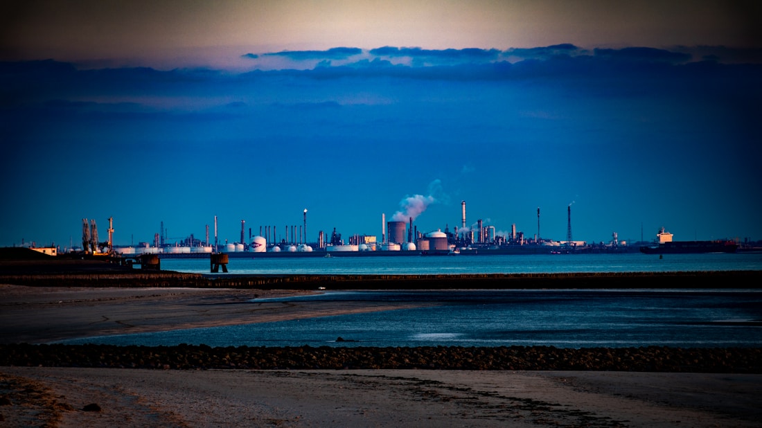 An industrial landscape featuring numerous large cylindrical tanks and distillation columns in the background, likely part of a refinery or chemical plant. Smoke is visible rising from some structures, indicating active industrial processes. The foreground includes a sandy shoreline with calm waters reflecting the blue and gray tones of the sky.