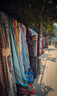 A person is arranging a colorful assortment of fabrics hanging vertically on a roadside stand. The fabrics include a variety of patterns and textures, such as checks and stripes. The setting is open-air with sunlight casting shadows on the concrete ground. A tree provides some shade, and a lamppost is seen nearby with various posters attached.